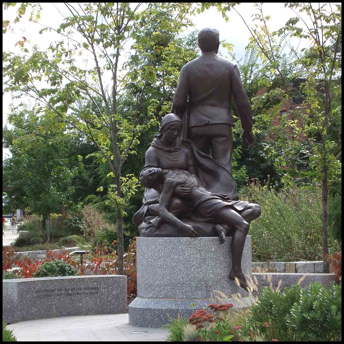 photo of bronze figural group on round stone base surrounded by pathway, shrubs, and trees