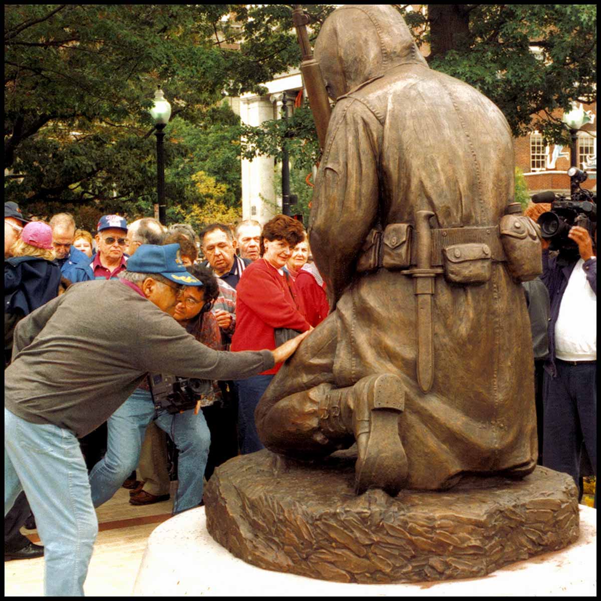 photo of man laying hand on bronze sculpture of kneeling Korean War soldier viewed from the back with crowd surrounding them and trees and buildings in background