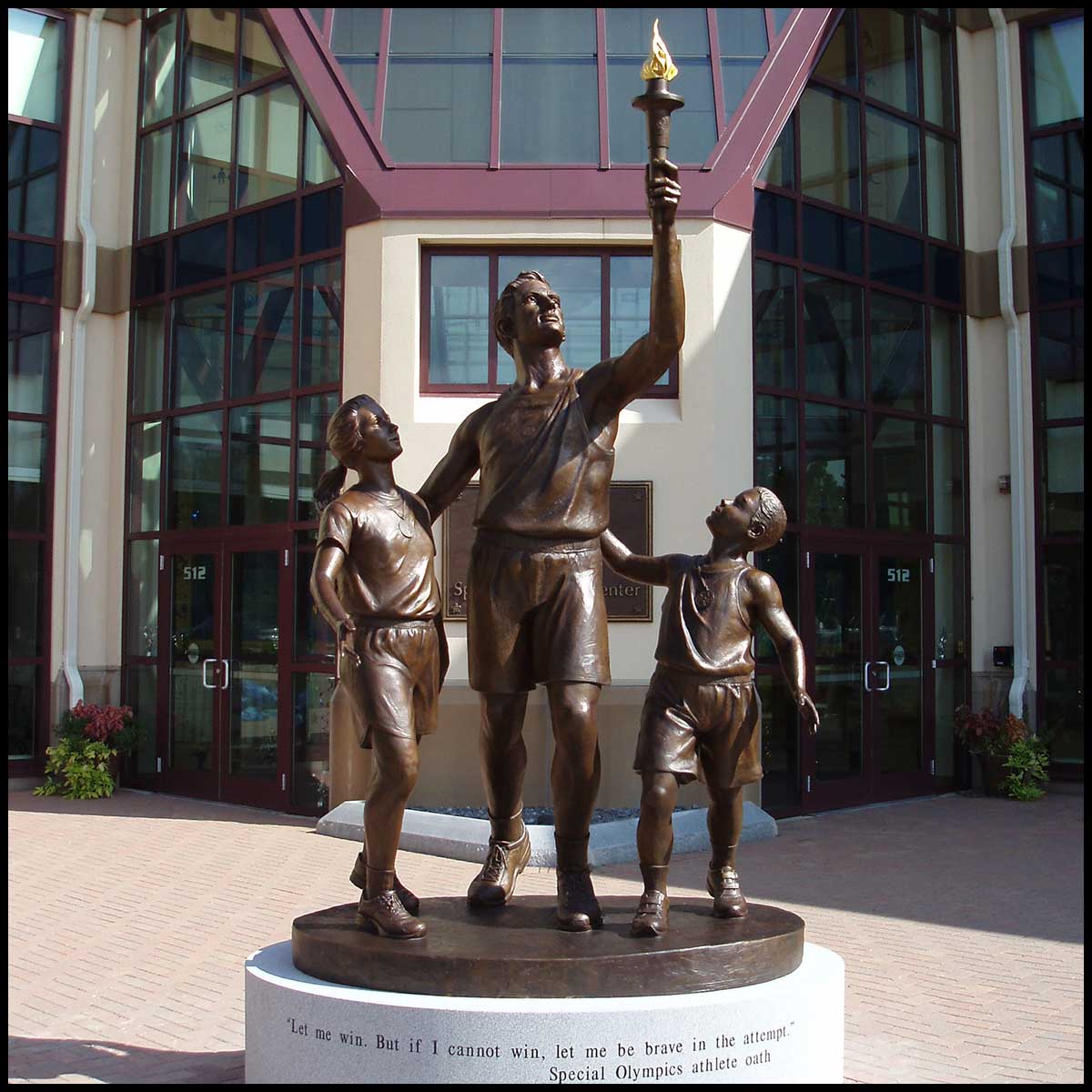 photo of bronze sculpture of man holding up Olympic torch with a boy and girl at his sides, placed on round stone base in plaza in front of building