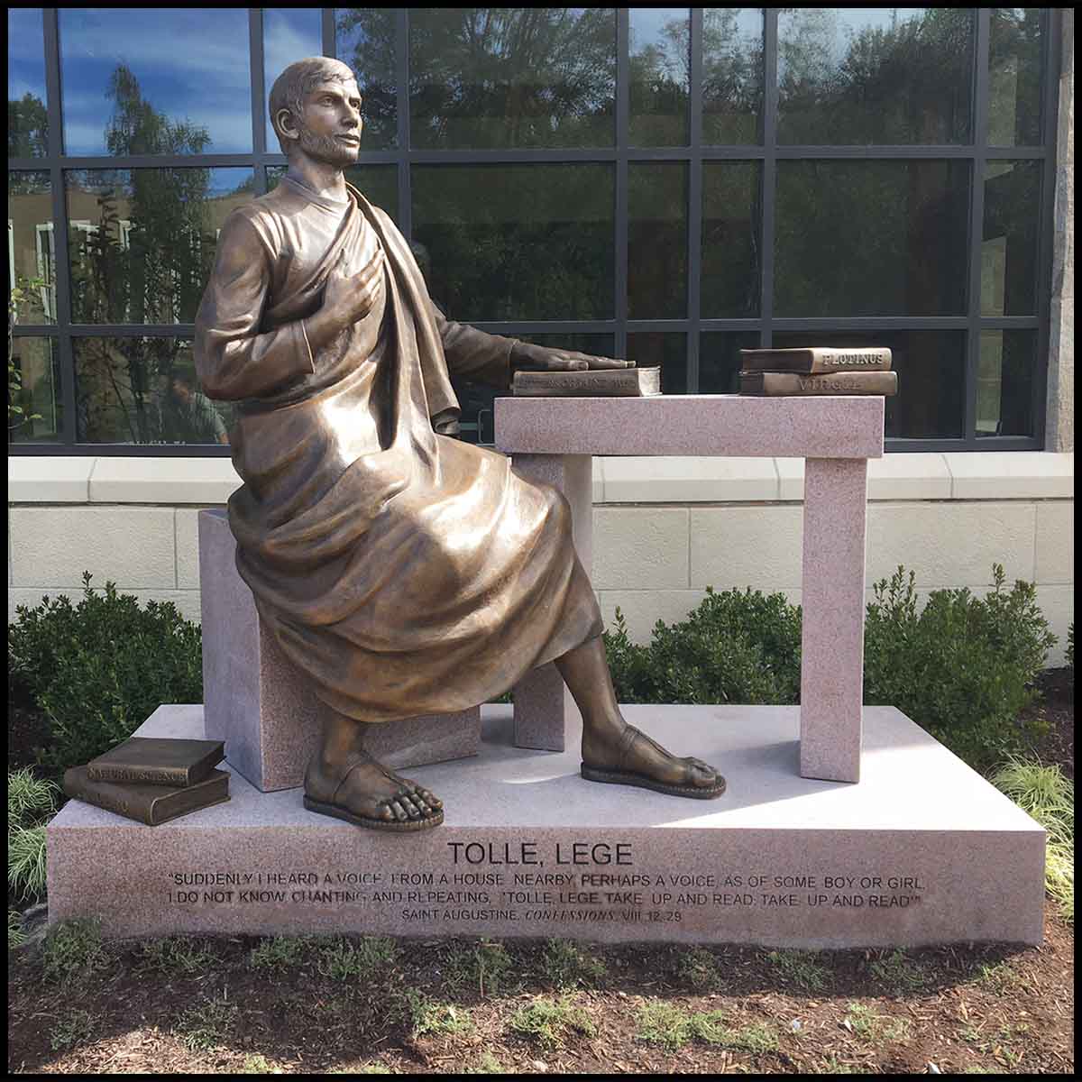 photo of bronze sculpture of man in robes seated on stone block with one hand on stone table and other raised to heart with windowed building behind