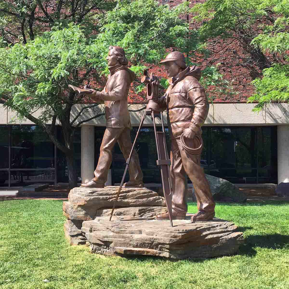photo of bronze sculpture of man and woman in winter gear with theodolite on tripod and woman taking notes, standing on rock surface in front of a building on a green lawn
