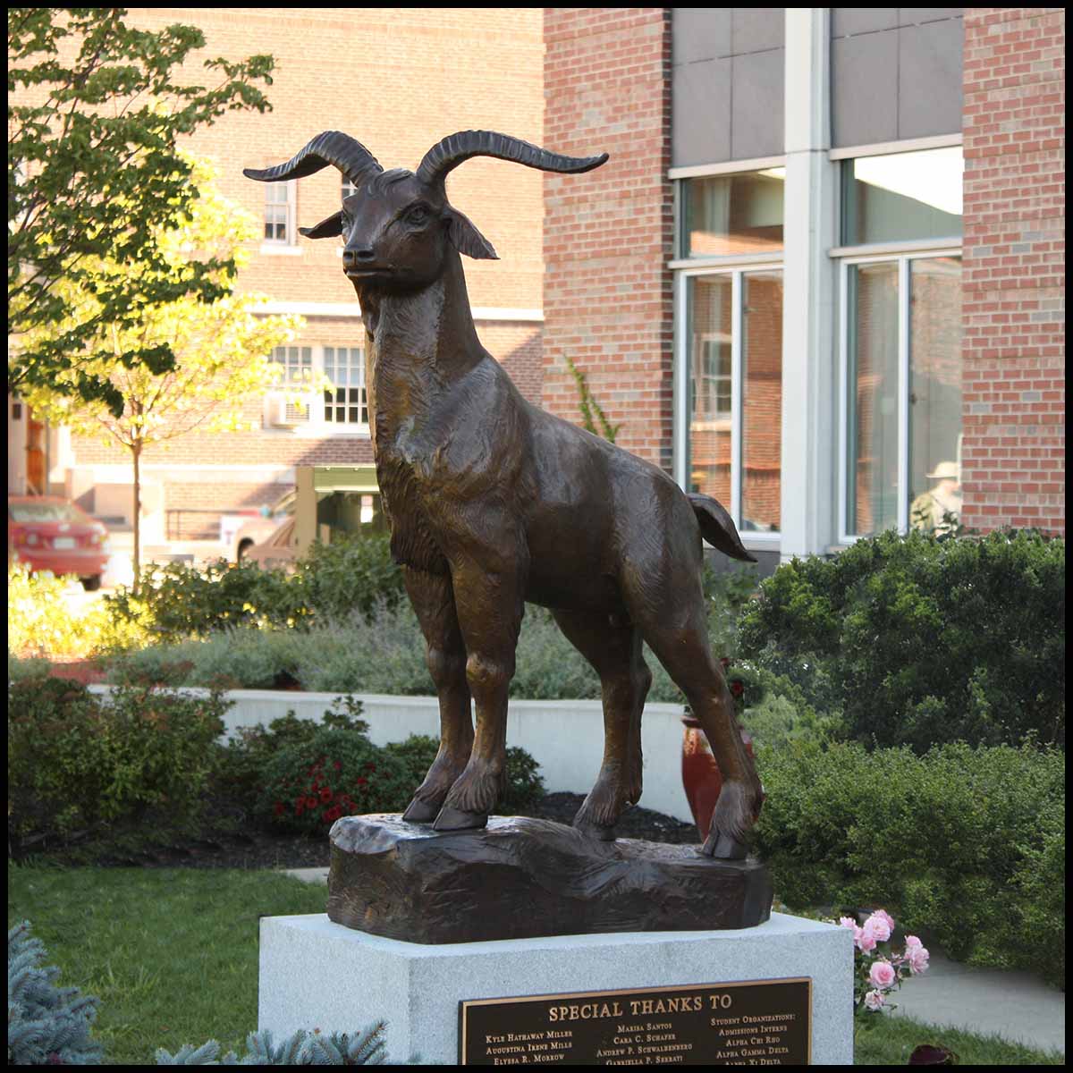 photo of bronze sculpture of goat standing on sculpted incline on granite base surrounded by greenery and brick buildings