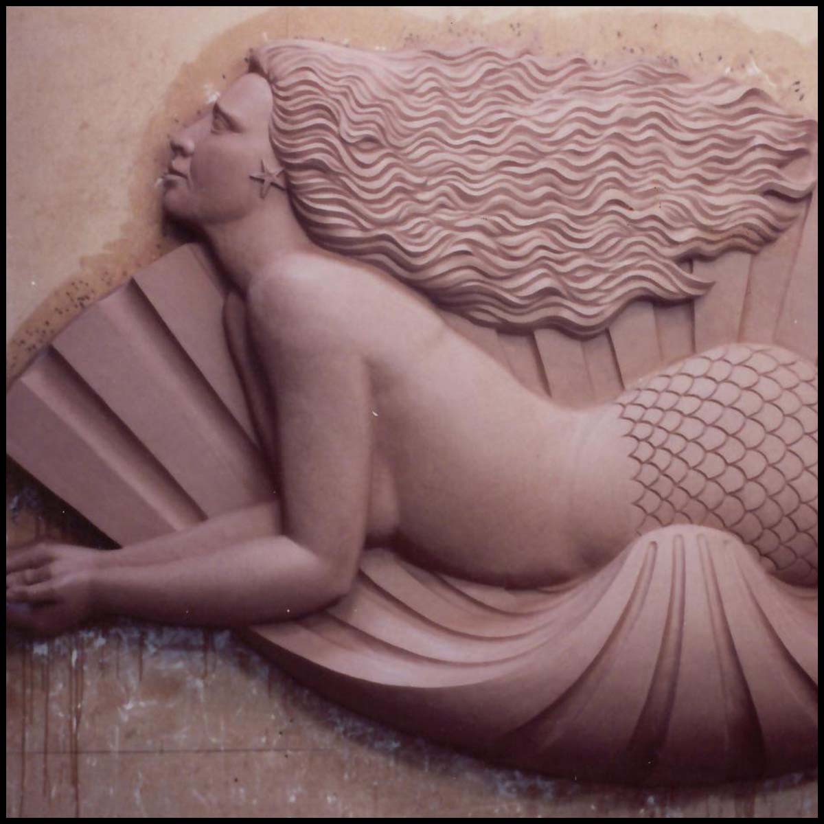photo of clay model of sculpture of mermaid in profile laying on a shell on wood backing leaning against wall