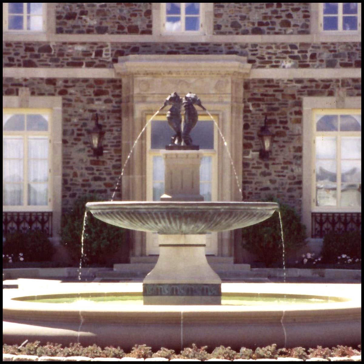 photo of granite fountain topped with bronze sculpture of two seahorses spouting water in front of a brick house