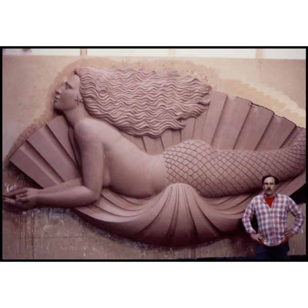 photo of clay model of sculpture of mermaid in profile laying on a shell on wood backing leaning against wall with sculptor Robert Shure standing in front